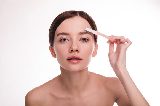 Portrait of beautiful slim and thin young woman look straight and tke care about eyebrows. Cut hair with small razor. Naked with glowing healthy skin. Isolated over white background.