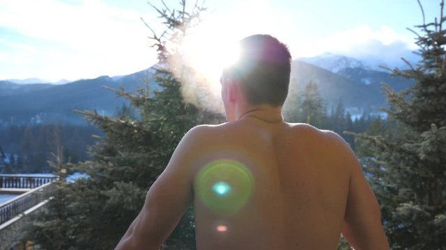 Rear view of young athletic guy stands on balcony, raises hands and stretches himself at sunny day. Handsome man yawns and covers her mouth breathing fresh air during winter morning. Slow motion
