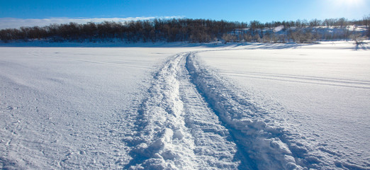 Trail from a snowmobile on a frozen and snowy river bed. Panoramic view of the river bank. Norway