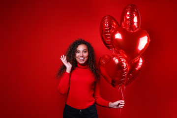 Studio portrait of young woman with dark skin and long curly hair wearing knitted turtle neck sweater over the festive red wall with heart shaped balloon. Close up, isolated background, copy space.