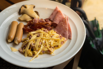 A set of breakfast that compose of scrambled eggs with cheese and ham, sausage, bacon, ham, and grilled mushroom placed on a white plate on a wooden table.