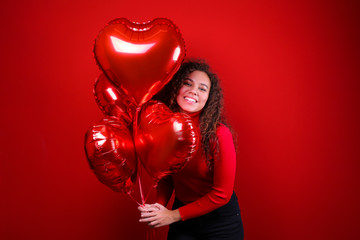 Studio portrait of young woman with dark skin and long curly hair wearing knitted turtle neck sweater over the festive red wall with heart shaped balloon. Close up, isolated background, copy space.