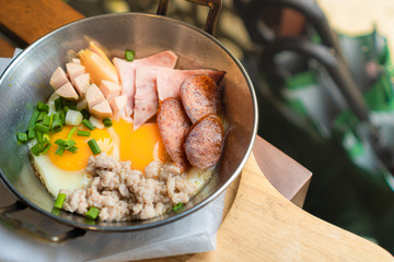 A breakfast that composes of fried egg, sausage, ham, and minced pork put in a small pan placed on wooden table