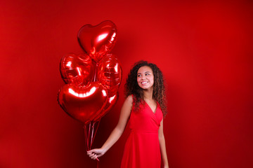 Studio portrait of young woman with dark skin and long curly hair wearing sexy dress over the festive red wall with heart shaped balloon. Close up, isolated background, copy space.