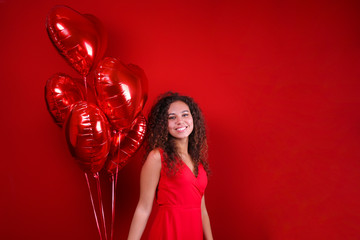 Studio portrait of young woman with dark skin and long curly hair wearing sexy dress over the festive red wall with heart shaped balloon. Close up, isolated background, copy space.