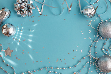 Bright festive background in silver and blue colors, flat lay. Frame of shiny Christmas balls with highlights, bows, serpentine, beads and star-shaped confetti, empty space for text in the center