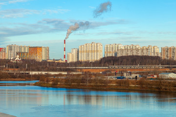 Fototapeta premium City landscape. In the foreground is a river. In the background are residential buildings, the boiler pipe from which comes smoke and a passing electric train.