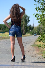 back shot of attractive brown-haired young woman worn in blue jeans shorts black top shod in high heel shoes standing on country road and waiting for a car	