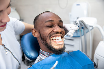 Fototapeta premium Young african-american man visiting dentist's office for prevention and treatment of the oral cavity. Man and woman doctor while checkup teeth. Healthy lifestyle, healthcare and medicine concept.