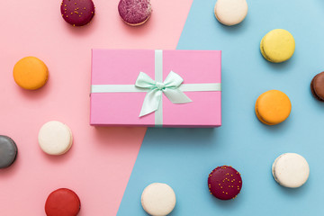 Multi-colored macaroon cakes on a pink and blue pastel background with a gift box flat lay
