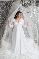 Preparing the bride for the upcoming wedding. Sexy bride. Bride's fees. Beautiful bride stands on a bed in a robe with a beautiful veil. Bride with luxurious hair and makeup. Wedding.  