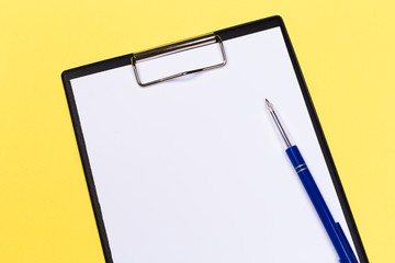 Clipboard with white sheet and pen on a yellow background. View from above. space for text