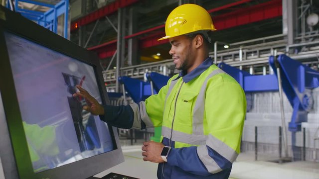 Black male engineer in uniform uses computer LCD touch screen for programming CNC machine or assembling components on conveyor. Work process on heavy industry plant or factory