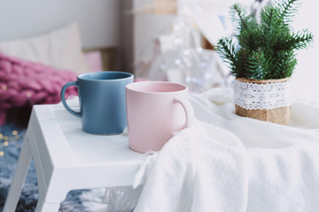 two pastel colored mugs on a small portable table by the window and Christmas decorations