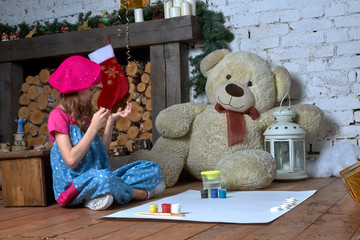 A little girl draws with her finger paints sitting on the floor. Christmas tree decorated with toys. Big Teddy bear. New year gift. Development. Childhood.Creativity. Hobby.
