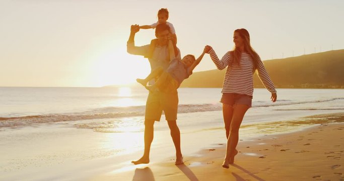 Two young toddler boys and their parents playing together happily on the beach at sunset, smiling parents swinging their son into the air, riding on dad's shoulders, happy family on vacation