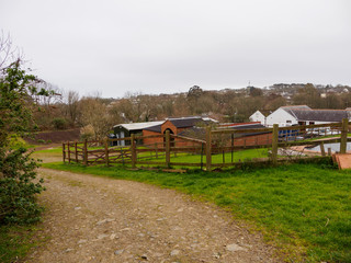 Wide angle view of a dirt road with wooden fences leading to old farmyard houses. Rosehill meadow, Penzance, United Kingdom. Agriculture and Cornish winemaking. - 310907344