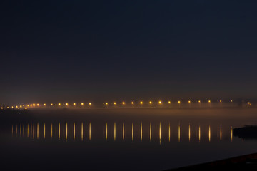 Obraz premium A bridge across the river lit by yellow lanterns in the fog and reflections of light in the water.