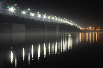 Night landscape of a road bridge over a river illuminated by colorful lights reflected in water and fog.