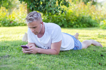 On a sunny day, a young man lies on the grass, looks into a smartphone, listens to music with wireless headphones. guy blond writes a message on the phone, checks social networks, plays a game.