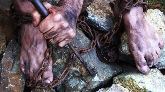 Man in captivity. A slave in iron chains is trying to free himself. A scene from the life of a slave. The symbol of slave labor. Hands and feet in chains. Breaking chains