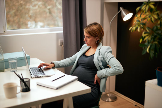 Cute caucasian pregnant businesswoman sitting on chair in office, holding back and using laptop.