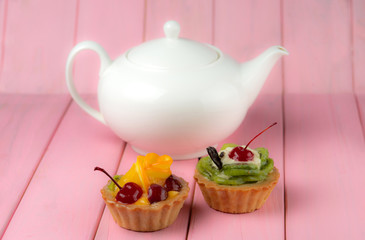 Colorful fruit cakes with kiwi, orange, candied cherries, cream and chocolate on a pink background with a teapot.