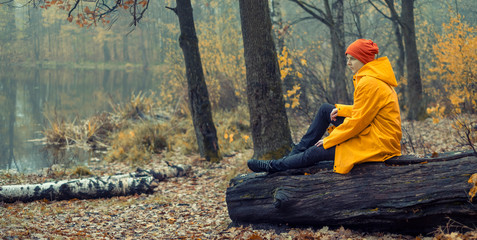young   girl in a yellow raincoat sits on a log on the shore of a fall forest lake alone and...