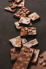 The chunks of broken different chocolate bars with nuts on the black stone surface with space for text. Background with chocolate. Sweet food photo concept.