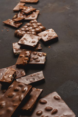 The chunks of broken different chocolate bars with nuts on the black stone surface with space for text. Background with chocolate. Sweet food photo concept.