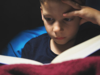 Little child, 7 years old boy reading a book inside at dim spot light