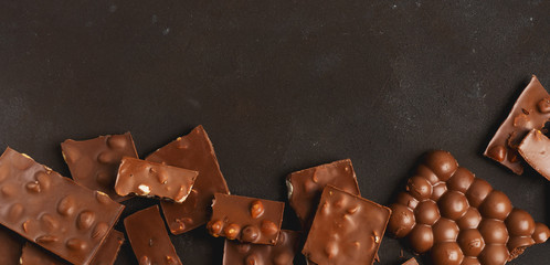 The chunks of broken different chocolate bars with nuts on the black stone surface with space for text. Background with chocolate. Sweet food photo concept.