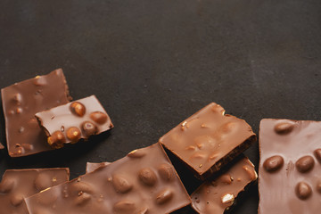 The chunks of broken different chocolate bars with nuts on the black stone surface with space for text. Background with chocolate. Sweet food photo concept.