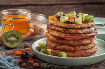 On the table is a dish with pancakes, honey, kiwi, nuts and raisins.