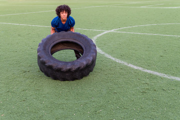 Latin fit young man with afro style hair in blue t-shirt training outdoors and flipping a tire during a crossfit WOD on a sunny day at the sports club.