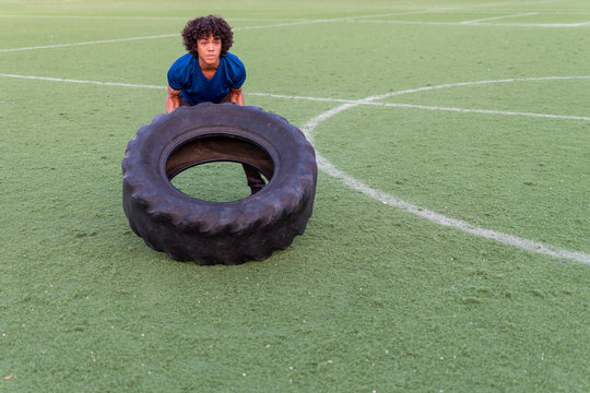 Latin fit young man with afro style hair in blue t-shirt training outdoors and flipping a tire during a crossfit WOD on a sunny day at the sports club.