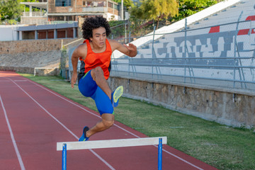 Obraz premium Latin fit young man with afro style hair in orange tank top at the running track jumping the hurdless fence on a sunny day.