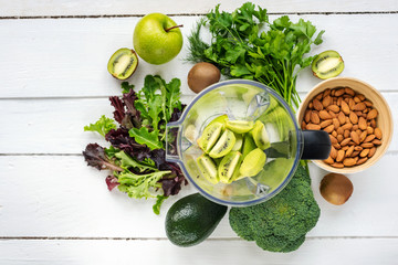 Ingredients for green detox smoothies in a blender: apples, kiwi, avocado, lettuce, broccoli, almonds,  dill and parsley on white wooden table. Top view.