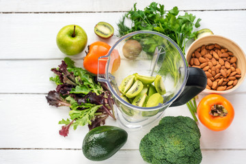 Ingredients for detox smoothies in a blender: apples, kiwi, avocado, persimmon, lettuce, broccoli, almonds,  dill and parsley on white wooden table. Top view.