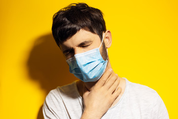 Portrait of young sick man wearing medical flu mask, holding neck with hand on yellow background.