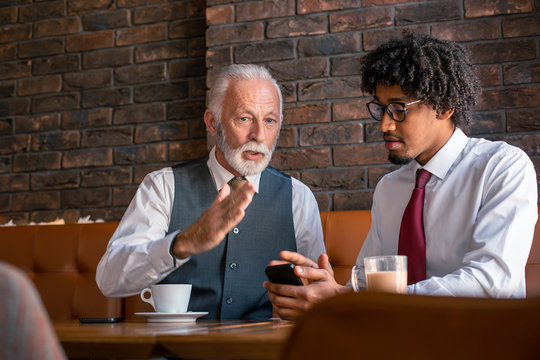An older executive explaining business plan to his younger employee while the employee is checking the data over the phone.