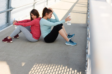 Two tired caucasian sports women in sweatshirts and leggings sitting on bridge back to back and resting after running.