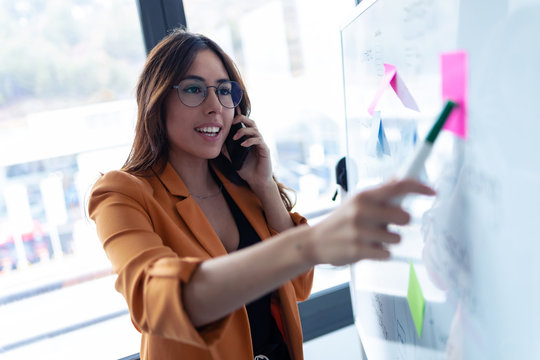 Business Young Woman Talking On Mobile Phone While Working On White Board With Post It Stickers In The Office.