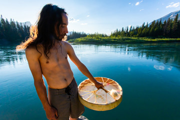 A slim and toned shirtless man wearing shorts is seen standing on the shore of a calm turquoise lake holding a sacred mystical drum 