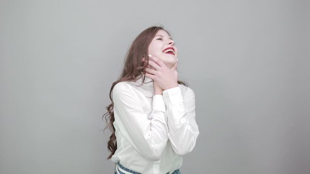 Attractive young woman in fashion white shirt holding hands on neck, choking herself, looking up isolated on gray background in studio. People sincere emotions, lifestyle concept.