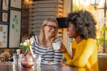 Two multi ethnic girl friends enjoying coffee together in a coffee shop, sitting at a table chatting and laughing loud.