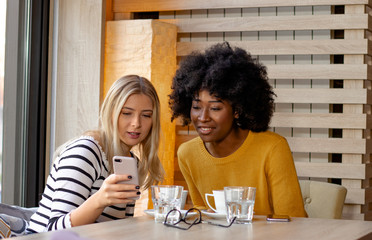 Two multi ethnic girl friends enjoying coffee together in a coffee shop, sitting at a table and looking pictures or social media content on a smartphone.