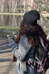 Fototapeta premium Brunette girl with curly hair checking mobile and looking to a lake while in a park during winter in London