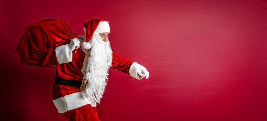 Portrait of crouching Santa Claus carrying huge red sack with presents on red studio background. Isolate.