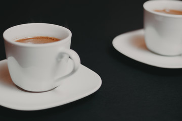 White color cup of coffee with saucer on black background, another one in blurred background